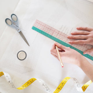 Dressmaking tools such as scissors, washi tape, measuring trape and clover ruler with a hand holding a pencil tracing a sewing patter throught patterntrace swedish tracing paper